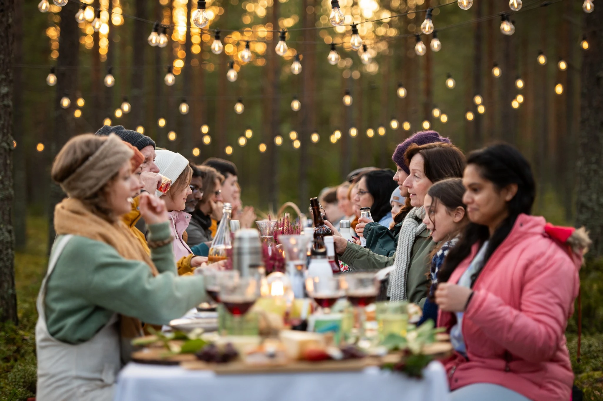 People eating at a table in a forest setting with string lights on the trees