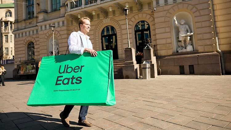 Mies kantaa Helsingissä Vanhan ylioppilastalon edessä suurta vihreää kassia jossa lukee Uber Eats