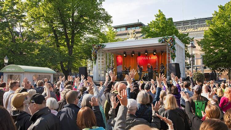 Espan lavan ulkoilmakonsertissa suuri yleisö nostaa kädet ilmaan esiintyjän soittaessa lavalla vehreässä puistoympäristössä.