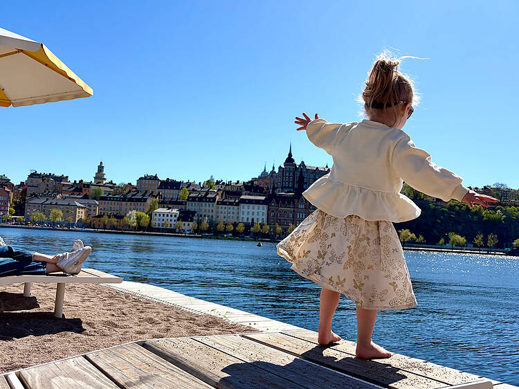 Young child balancing on a waterside deck in Stockholm on a sunny day, with Riddarholmen’s skyline and blue water in the background. Young child balancing on a waterside deck in Stockholm on a sunny day, with Riddarholmen’s skyline and blue water in the background.