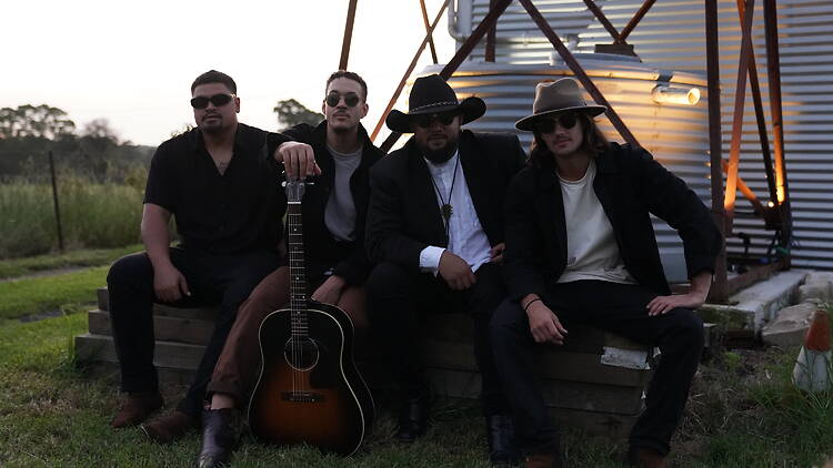 Simply West A group of four men sitting outside with a guitar