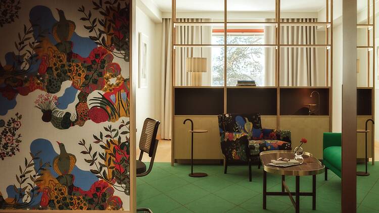 Moderni hotellihuone, jossa värikäs kuviollinen seinä, avonainen puinen hyllyjakaja ja design-nojatuoli muodostavat viihtyisän oleskelutilan pehmeässä valaistuksessa.