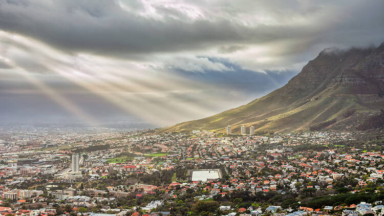 Sunlight beams breaking through dark storm clouds over Cape Town suburbs Cape Town at the foot of Table Mountain, steep ,Sunbeams pierce through clouds, illuminating the urban landscape and residential neighborhoods of, grassy slopes.