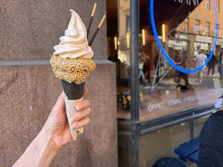 Soft serve ice cream cone in Stockholm, where the subject in the image is a creamy swirl topped with crunchy coating and chocolate sticks, held in front of a gelato shop window. Soft serve ice cream cone in Stockholm, where the subject in the image is a creamy swirl topped with crunchy coating and chocolate sticks, held in front of a gelato shop window.