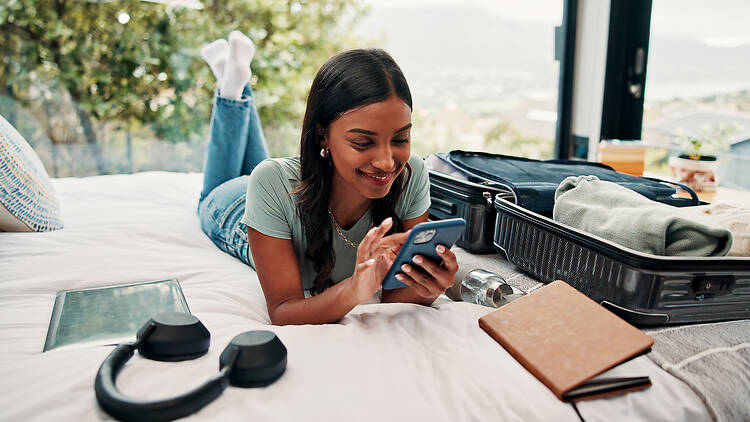 2248369530 Relax, phone and luggage with woman in bedroom for travel blog, social media and holiday post. Online booking, trip research and resort review with person and mobile in house rental for contact