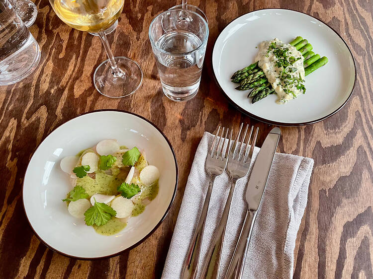 Stilren restaurangservering i Malmö med sparris i krämig sås, förrätt med örter och skivad fisk samt vitt vin och vatten på ett träbord. Stilren restaurangservering i Malmö med sparris i krämig sås, förrätt med örter och skivad fisk samt vitt vin och vatten på ett träbord.