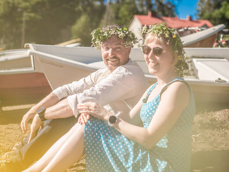 Kaksi kukkaseppeleisiin pukeutunutta henkilöä istuu aurinkoisella nurmella veneiden vieressä kesäisessä järvimaisemassa. Kaksi kukkaseppeleisiin pukeutunutta henkilöä istuu aurinkoisella nurmella veneiden vieressä kesäisessä järvimaisemassa.