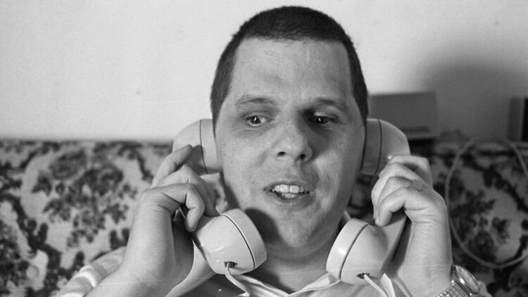  Joybubbles ( Rachael J. Morrison , 2026 ) Image description: Black - and - white film still showing a man seated on a patterned sofa holding two telephone receivers to his ears.