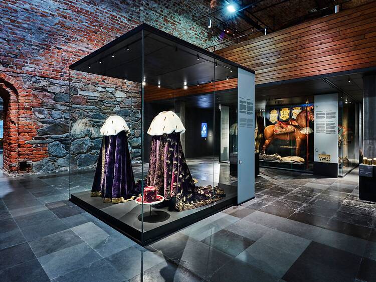 En museisal med tegelväggar och glasmontrar visar historiska kungliga dräkter och föremål, där en rikt dekorerad häst och rustning syns i bakgrunden. En museisal med tegelväggar och glasmontrar visar historiska kungliga dräkter och föremål, där en rikt dekorerad häst och rustning syns i bakgrunden.