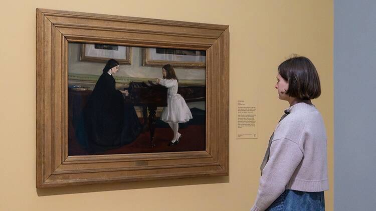 A woman looks at ‘At The Piano’, a portrait of a woman playing piano and a girl dressed in white listening