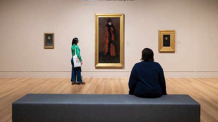 A woman sits on a bench in front of three Whistler paintings, with another woman stood in front of the central painting