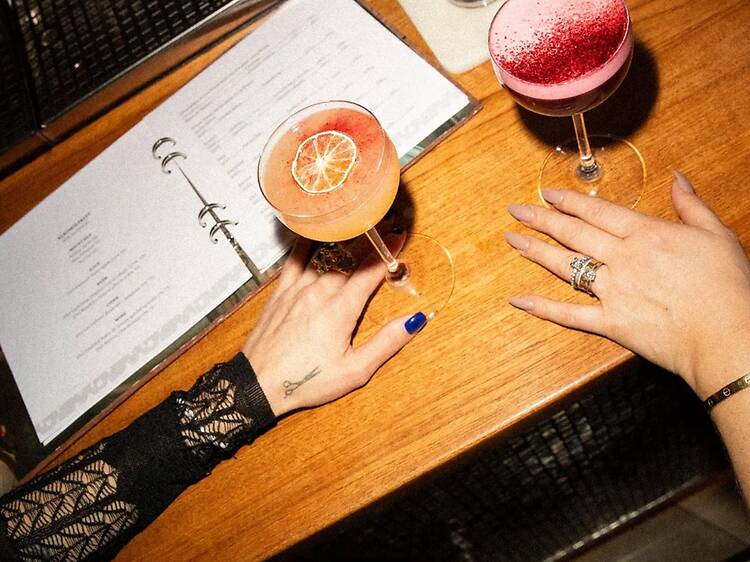 Two cocktails and a menu rest on a wooden table at Avalon bar in Gothenburg, with hands reaching for the drinks. Two cocktails and a menu rest on a wooden table at Avalon bar in Gothenburg, with hands reaching for the drinks.