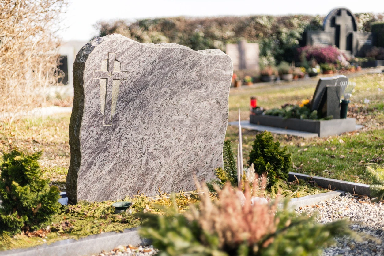 A grave marker stands in a cemetery with green trees and flower arrangements nearby. The sun shines on the stones, highlighting the surroundings. The scene shows care and remembrance.