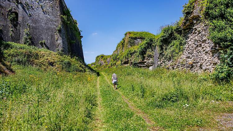 Trail between massive high stone walls at Charlemont fortress against blue sky in background, female tourist walking with back to camera, sunny summer day in Givet, Ardennes department, France