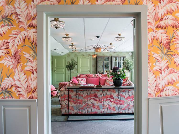 An interior view framed by a doorway looking into a brightly decorated lounge room, featuring vibrant orange and pink botanical wallpaper An interior view framed by a doorway looking into a brightly decorated lounge room, featuring vibrant orange and pink botanical wallpaper