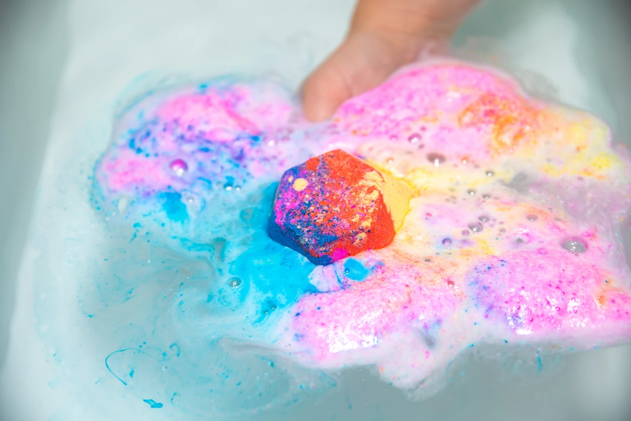 Tie-dye colored (red, orange, blue, pink and yellow) bath bomb with multi-colored bubbles partially submerged in water with a hand holding it