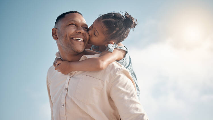 Smiling mixed race single father carrying little daughter on piggyback with copyspace. Adorable, happy, hispanic girl bonding with parent and kissing cheek on beach. Man and child enjoying free time
