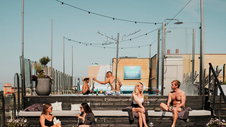 Selma City Spa, Clarion Sign People relaxing and chatting on cushioned wooden bleachers at a sunny rooftop pool lounge under string lights.