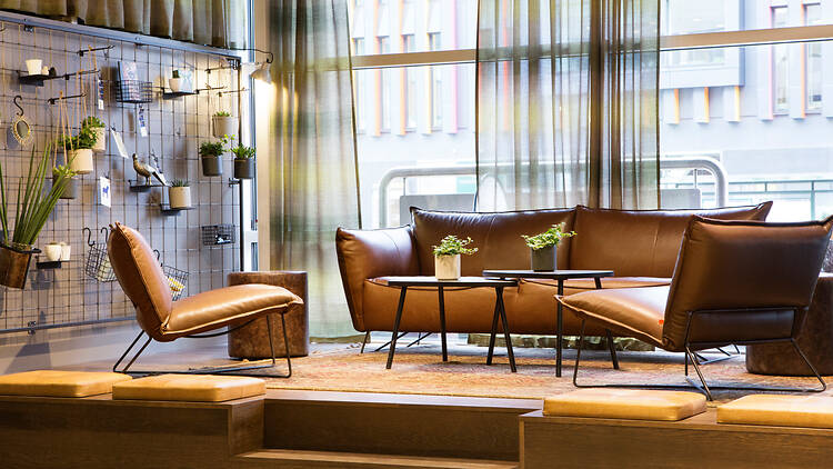Comfort Hotel Xpress Lobby, Strawberry A stylish hotel lobby lounge featuring brown leather seating, a raised wooden platform, and large windows with sheer curtains.