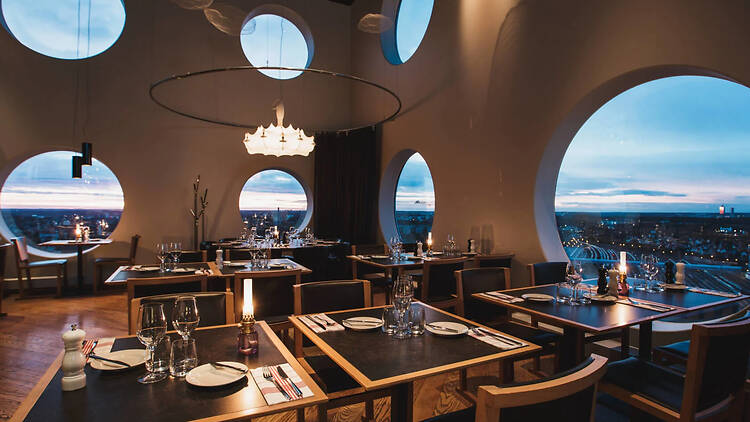 Quality Hotel Strawberry Arena, Strawberry An elegant hotel restaurant featuring set dining tables and distinctive, large circular windows overlooking a city view at dusk.