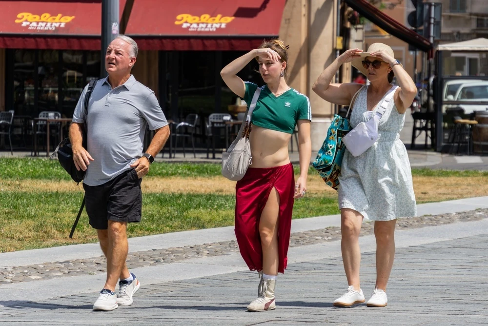 Three tourists walking in city square shielding eyes from intense summer sun