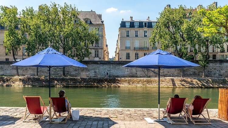 People relaxing on red deckchairs under blue umbrellas on the riverbank of the Seine on a hot summer's day in Paris People relaxing on red deckchairs under blue umbrellas on the riverbank of the Seine on a hot summer's day in Paris