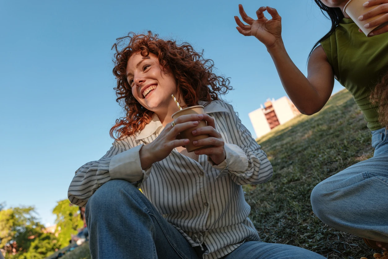 Two young friends share a casual moment on a sunny park day in Madrid, sipping drinks and laughing. The relaxed scene highlights friendship, connection, and outdoor leisure in a bright urban setting.