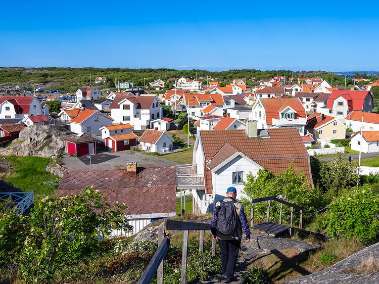 A hiker with a backpack walks down a wooden staircase overlooking a dense cluster of white and red houses on the island of Vrångö under a bright blue sky. A hiker with a backpack walks down a wooden staircase overlooking a dense cluster of white and red houses on the island of Vrångö under a bright blue sky.