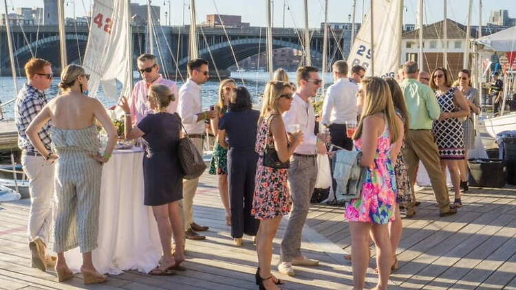 A wide shot of people enjoying drinks at a marina with sailboats in the background. A wide shot of people enjoying drinks at a marina with sailboats in the background.