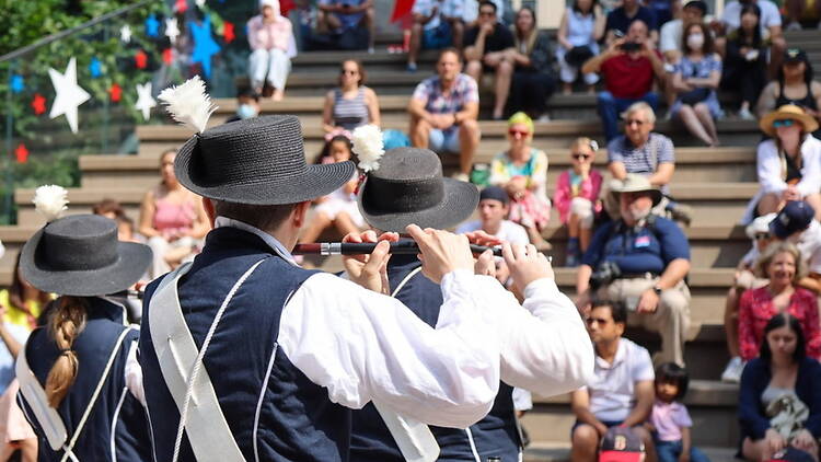 A wide shot of a performer playing a flute in front of an audience in an outdoor park. A wide shot of a performer playing a flute in front of an audience in an outdoor park.