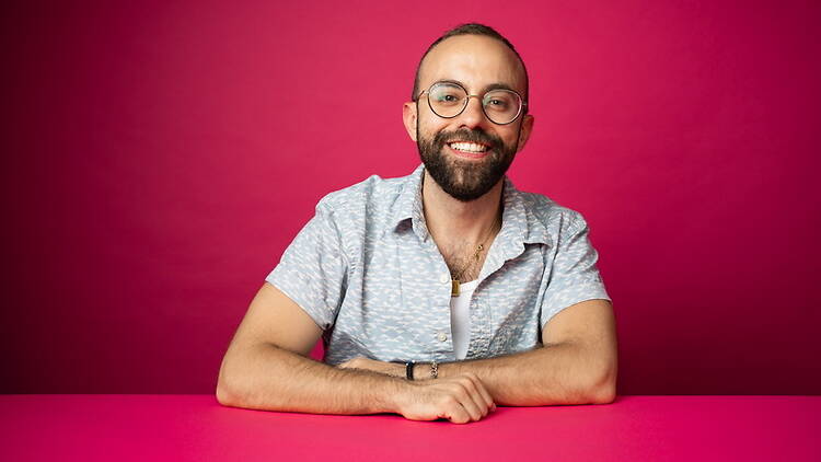 Medium shot of a smiling person sitting at a table against a bright pink background. Medium shot of a smiling person sitting at a table against a bright pink background.
