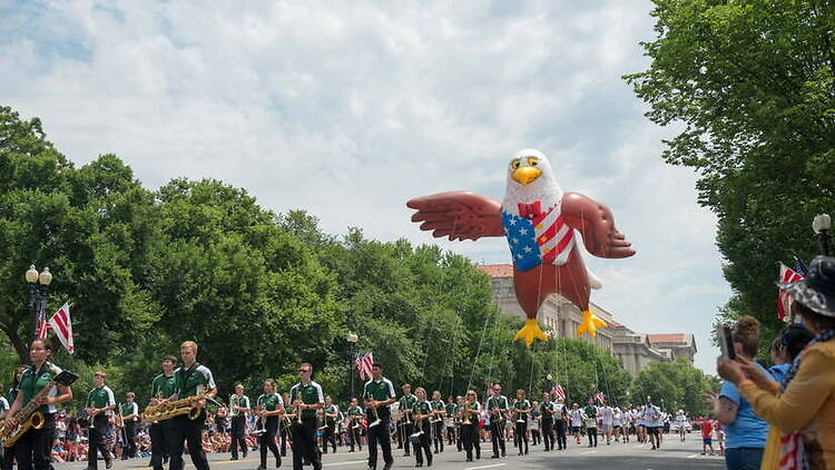 Wide shot of a marching band performing down a street alongside a large, patriotic bald eagle balloon during a daytime parade Wide shot of a marching band performing down a street alongside a large, patriotic bald eagle balloon during a daytime parade