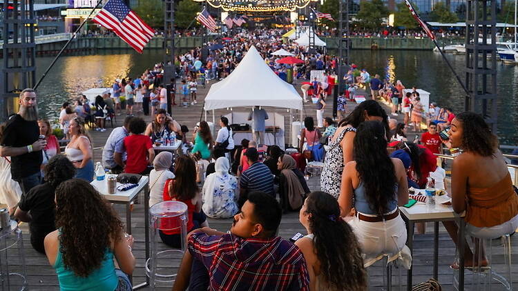 Wide shot of a large crowd of people socializing and dining on a waterfront pier decorated with American flags Wide shot of a large crowd of people socializing and dining on a waterfront pier decorated with American flags