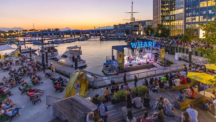 Wide shot of a crowd of people sitting outdoors and watching a band perform on a floating stage labeled "WHARF" at a waterfront harbor Wide shot of a crowd of people sitting outdoors and watching a band perform on a floating stage labeled "WHARF" at a waterfront harbor