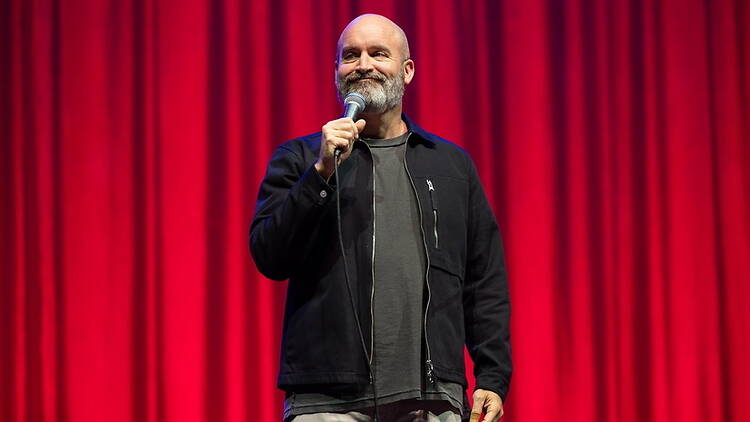 A close-up of Tom Segura holding a microphone on stage. A close-up of Tom Segura holding a microphone on stage.