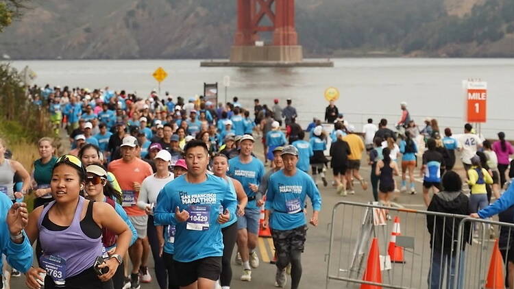 Wide shot of runners participating in a marathon along a waterfront beneath a large suspension bridge.