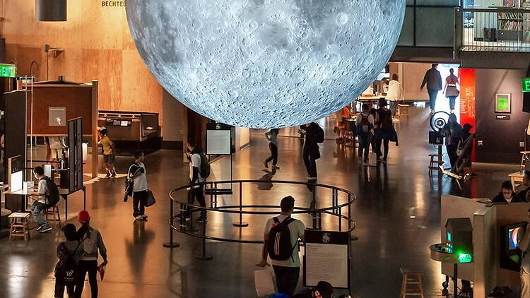 Wide shot of people exploring exhibits beneath a large suspended moon model in a science museum Wide shot of people exploring exhibits beneath a large suspended moon model in a science museum