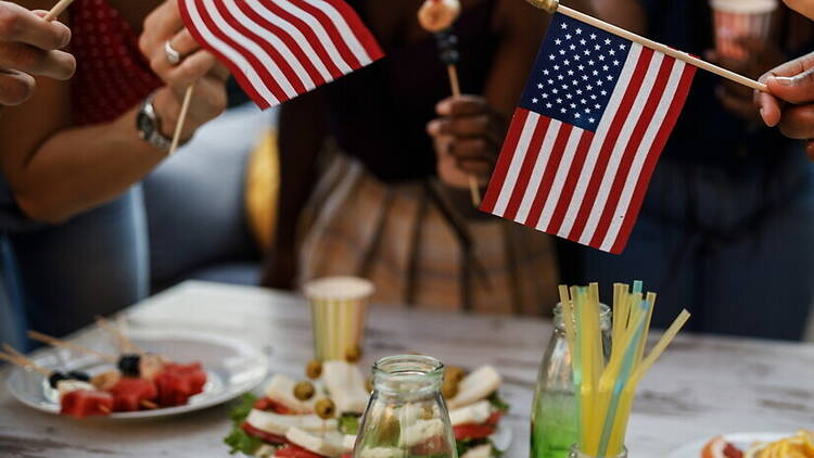 Close-up view of people holding small American flags over a table filled with party food and drinks Close-up view of people holding small American flags over a table filled with party food and drinks