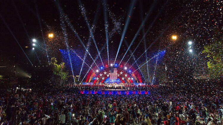 A wide shot of people watching a live performance as confetti falls at an outdoor event. A wide shot of people watching a live performance as confetti falls at an outdoor event.