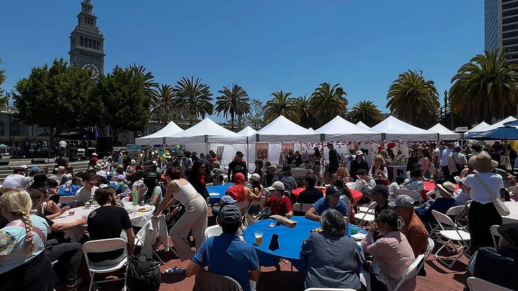 Wide shot of a large crowd sitting at tables and exploring market stalls in an outdoor plaza filled with white tents and palm trees beneath a clock tower Wide shot of a large crowd sitting at tables and exploring market stalls in an outdoor plaza filled with white tents and palm trees beneath a clock tower