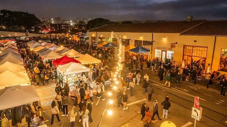 Wide shot of a large crowd walking and shopping at an outdoor night market illuminated by string lights Wide shot of a large crowd walking and shopping at an outdoor night market illuminated by string lights