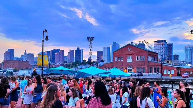 A wide shot of people gathering at a waterfront venue with a city skyline A wide shot of people gathering at a waterfront venue with a city skyline