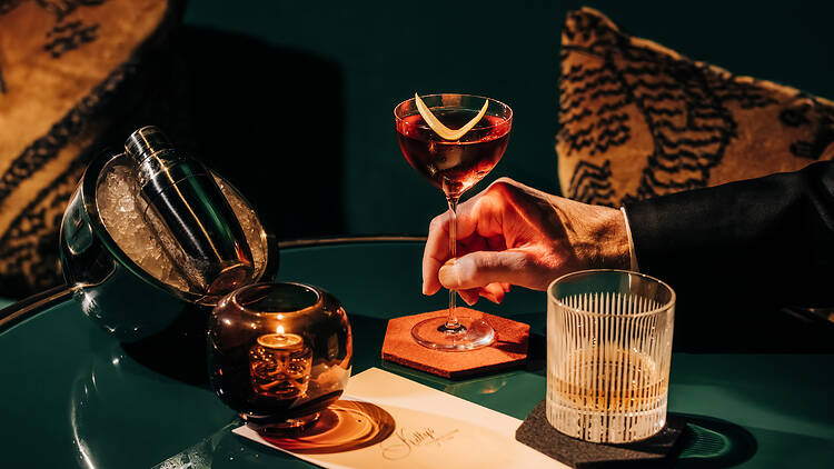 An arm holding a glass on a table alongside two other drinks and a shaker over a bed of ice.