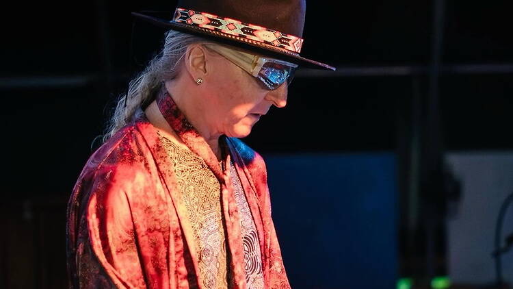 Medium close-up of a person wearing a hat and sunglasses performing on a dimly lit stage. Medium close-up of a person wearing a hat and sunglasses performing on a dimly lit stage.