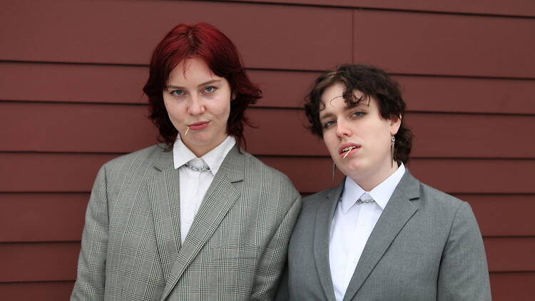 Medium shot of two people posing in matching suits against a paneled wall. Medium shot of two people posing in matching suits against a paneled wall.