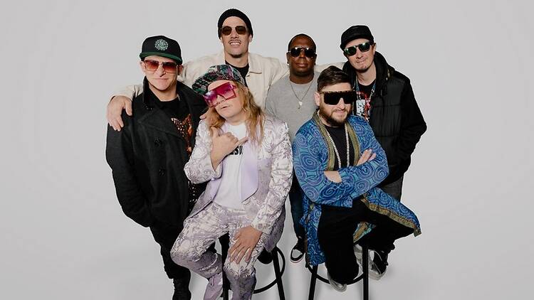 Wide shot of a six-member band posing against a plain white studio background.