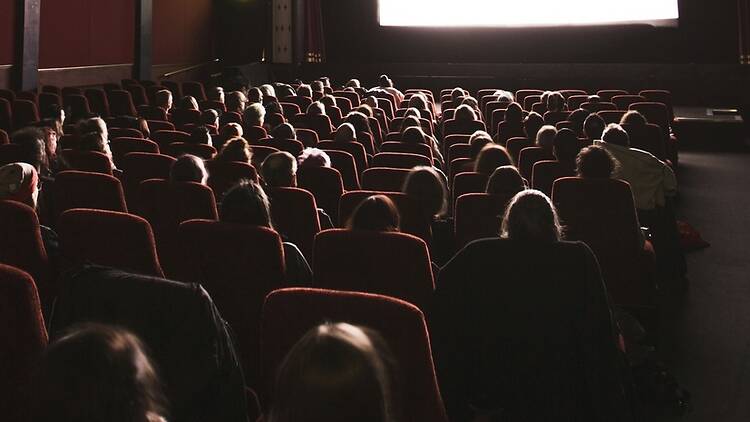 Wide shot rear view of an audience sitting in rows of red theater seats watching a movie screen in a dim cinema. Wide shot rear view of an audience sitting in rows of red theater seats watching a movie screen in a dim cinema.