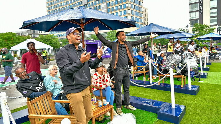 Wide shot view of people cheering at an outdoor viewing area under umbrellas Wide shot view of people cheering at an outdoor viewing area under umbrellas