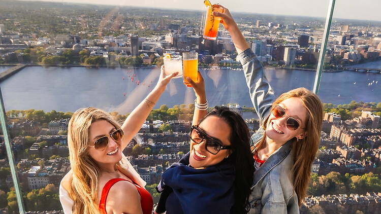 High-angle view of three women raising drinks above a city skyline. High-angle view of three women raising drinks above a city skyline.