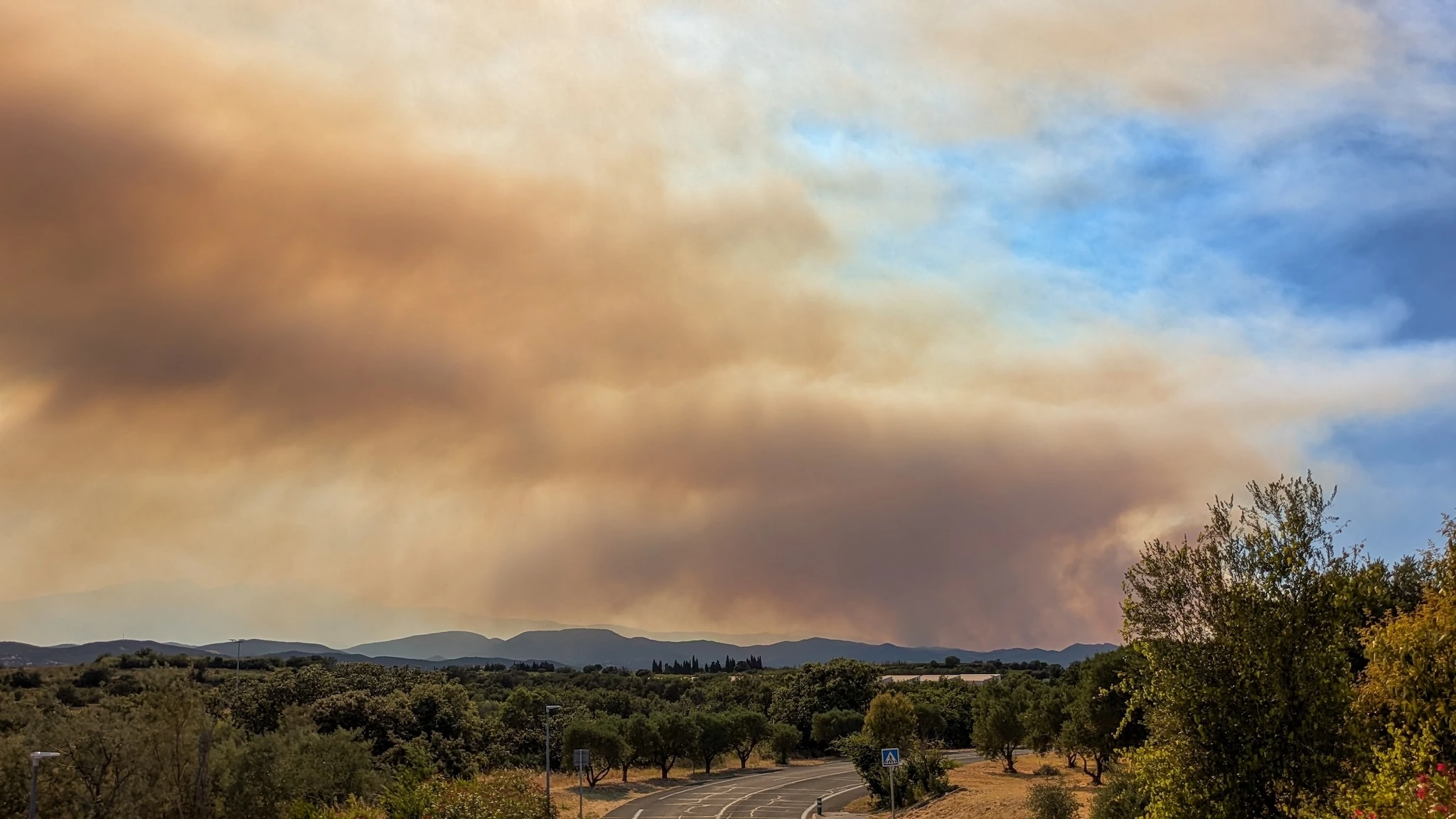Smoke from the Tr&eacute;villach wildfire spreads over a rural road and Mediterranean hills near Perpignan during a major wildfire in southern France.